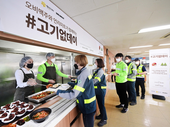 오비맥주 구매·지속가능경영 부문 나탈리 보르헤스 부사장과 김중수 전무가 오비맥주 이천공장 직원식당에서 직원들에게 식물성 대체육으로 만든 제육볶음, 카스 맥주박을 업사이클링해 만든 미니 초코링 등 저탄소 메뉴를 배식하고 있다. (사진=오비맥주)